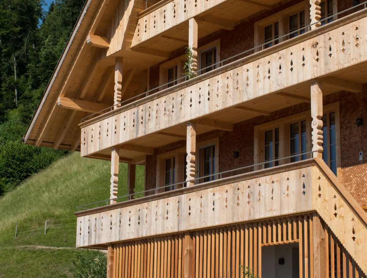 Mehrstöckiges Holzhaus im traditionellen Schweizer Stil mit kunstvoll verzierten Holzbalkonen und grosszügigem Dachüberstand. Die Fassade besteht aus hellem, massivem Holz, umgeben von einer grünen Hanglandschaft bei klarem Himmel.