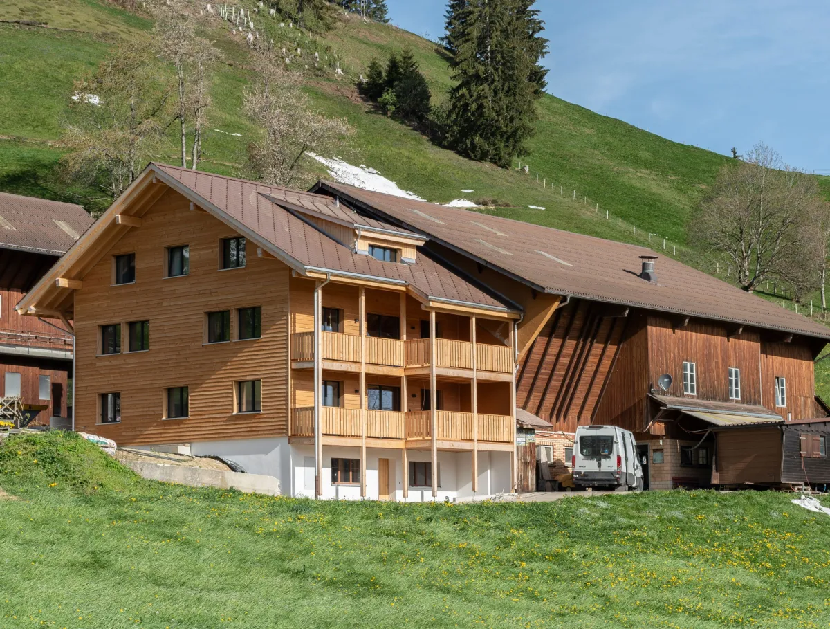 Aussenansicht eines neu errichteten Wohnhauses in Holzbauweise mit Satteldach und Balkonen, eingebettet in eine hügelige, ländliche Landschaft. Das Gebäude steht neben bestehenden landwirtschaftlichen Bauten. Im Vordergrund eine Wiese, im Hintergrund ein begrünter Hang mit Bäumen unter blauem Himmel.