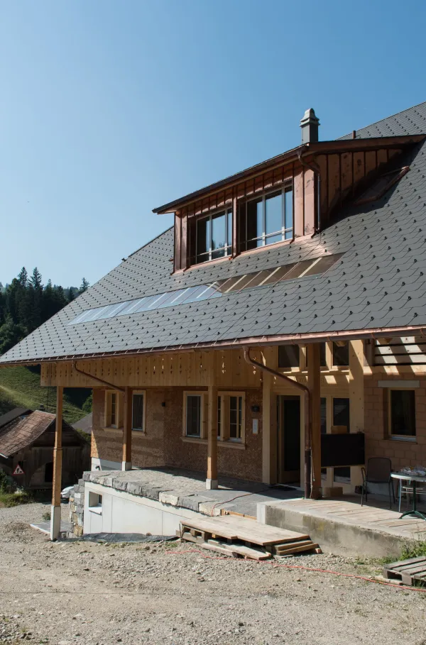 Mehrstöckiges Holzhaus im traditionellen Schweizer Stil mit kunstvoll verzierten Holzbalkonen und grosszügigem Dachüberstand. Die Fassade besteht aus hellem, massivem Holz, umgeben von einer grünen Hanglandschaft bei klarem Himmel.