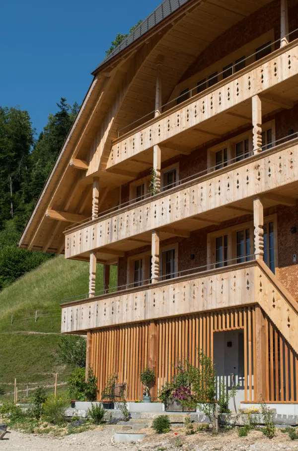 Mehrstöckiges Holzhaus im traditionellen Schweizer Stil mit kunstvoll verzierten Holzbalkonen und grosszügigem Dachüberstand. Die Fassade besteht aus hellem, massivem Holz, umgeben von einer grünen Hanglandschaft bei klarem Himmel.