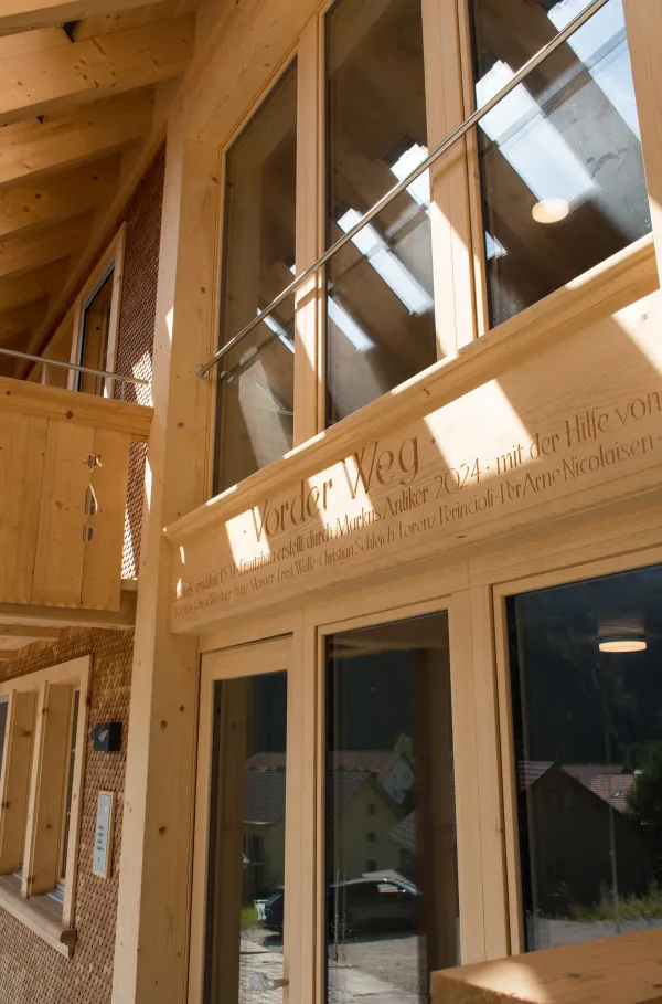 Mehrstöckiges Holzhaus im traditionellen Schweizer Stil mit kunstvoll verzierten Holzbalkonen und grosszügigem Dachüberstand. Die Fassade besteht aus hellem, massivem Holz, umgeben von einer grünen Hanglandschaft bei klarem Himmel.