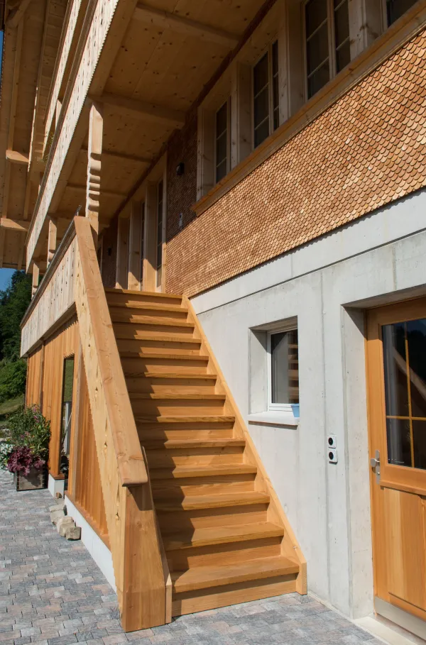 Mehrstöckiges Holzhaus im traditionellen Schweizer Stil mit kunstvoll verzierten Holzbalkonen und grosszügigem Dachüberstand. Die Fassade besteht aus hellem, massivem Holz, umgeben von einer grünen Hanglandschaft bei klarem Himmel.