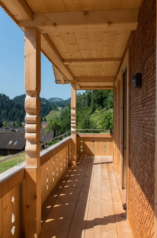 Mehrstöckiges Holzhaus im traditionellen Schweizer Stil mit kunstvoll verzierten Holzbalkonen und grosszügigem Dachüberstand. Die Fassade besteht aus hellem, massivem Holz, umgeben von einer grünen Hanglandschaft bei klarem Himmel.