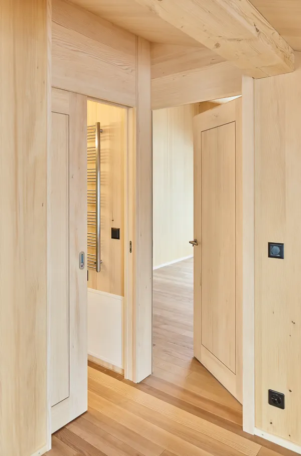 Innenaufnahme eines grosszügigen, lichtdurchfluteten Wohnraums in moderner Holzbauweise. Wände, Decke und Einbauten bestehen aus hellen Holzelementen und erzeugen eine warme, natürliche Atmosphäre. Grosse bodentiefe Fenster eröffnen den Blick auf verschneite Vegetation. Rechts liegt eine offene Küche mit Holzfronten und integrierten Schränken. Der durchgehende Holzboden verbindet alle Bereiche zu einem harmonischen Gesamtbild.
