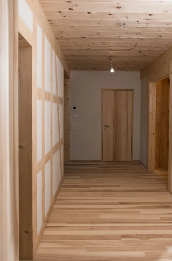 Innenaufnahme eines neu ausgebauten Dachraums mit sichtbarer Holzkonstruktion. Wände, Decke und tragende Balken bestehen aus hellen, sauber verarbeiteten Holzelementen, die eine warme und natürliche Atmosphäre schaffen. Der Raum verfügt über zwei Fenster, durch die Tageslicht einfällt und Schattenlinien auf die Wände wirft. Die Dachschräge und die präzise ausgeführten Holzverbindungen betonen die handwerkliche Qualität. Der Boden ist aus hellem, mehrfarbig geöltem Holz und verleiht dem Raum ein harmonisches