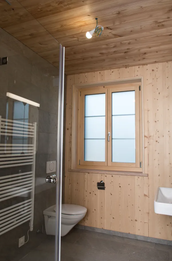 Innenaufnahme eines neu ausgebauten Dachraums mit sichtbarer Holzkonstruktion. Wände, Decke und tragende Balken bestehen aus hellen, sauber verarbeiteten Holzelementen, die eine warme und natürliche Atmosphäre schaffen. Der Raum verfügt über zwei Fenster, durch die Tageslicht einfällt und Schattenlinien auf die Wände wirft. Die Dachschräge und die präzise ausgeführten Holzverbindungen betonen die handwerkliche Qualität. Der Boden ist aus hellem, mehrfarbig geöltem Holz und verleiht dem Raum ein harmonisches