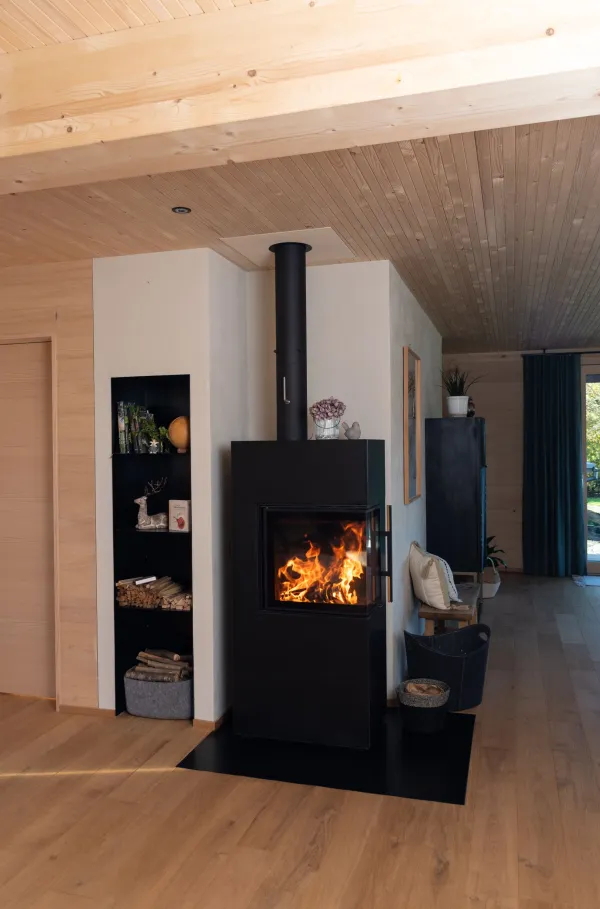 Innenansicht eines gemütlichen Wohnzimmers in moderner Holzbauweise. Wände und Decke sind mit hellem Holz verkleidet und schaffen eine warme Atmosphäre. Ein dunkles Sofa mit Kissen und Decke steht neben einem Sessel und einem kleinen Holztisch auf einem Teppich. Pflanzen, Steh- und Tischlampen sowie Tageslicht durch mehrere Fenster verleihen dem Raum eine wohnliche und einladende Stimmung.