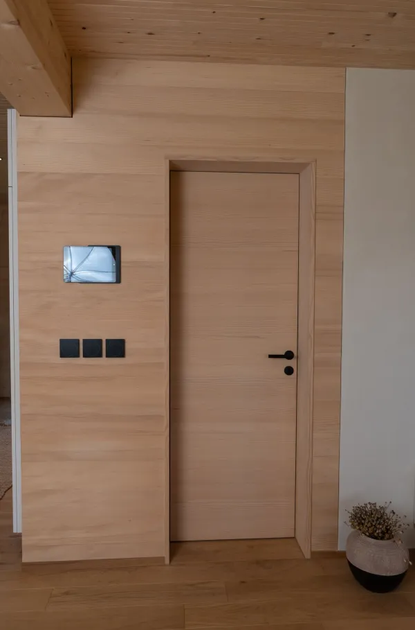 Innenansicht eines gemütlichen Wohnzimmers in moderner Holzbauweise. Wände und Decke sind mit hellem Holz verkleidet und schaffen eine warme Atmosphäre. Ein dunkles Sofa mit Kissen und Decke steht neben einem Sessel und einem kleinen Holztisch auf einem Teppich. Pflanzen, Steh- und Tischlampen sowie Tageslicht durch mehrere Fenster verleihen dem Raum eine wohnliche und einladende Stimmung.
