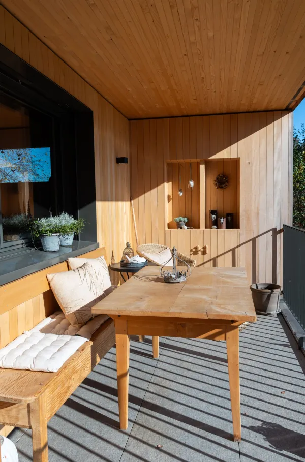 Innenansicht eines gemütlichen Wohnzimmers in moderner Holzbauweise. Wände und Decke sind mit hellem Holz verkleidet und schaffen eine warme Atmosphäre. Ein dunkles Sofa mit Kissen und Decke steht neben einem Sessel und einem kleinen Holztisch auf einem Teppich. Pflanzen, Steh- und Tischlampen sowie Tageslicht durch mehrere Fenster verleihen dem Raum eine wohnliche und einladende Stimmung.