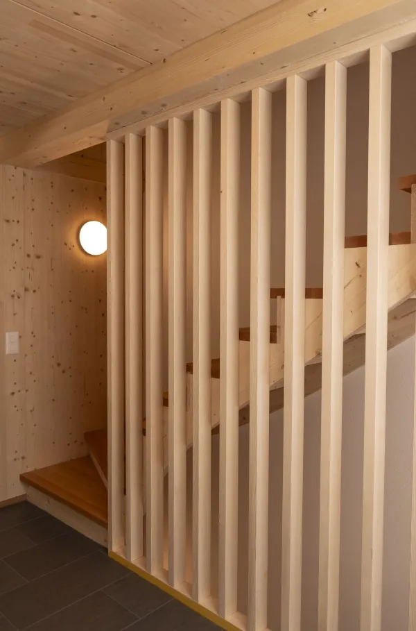 Aussenansicht eines neu errichteten Wohnhauses in Holzbauweise mit Satteldach und Balkonen, eingebettet in eine hügelige, ländliche Landschaft. Das Gebäude steht neben bestehenden landwirtschaftlichen Bauten. Im Vordergrund eine Wiese, im Hintergrund ein begrünter Hang mit Bäumen unter blauem Himmel.