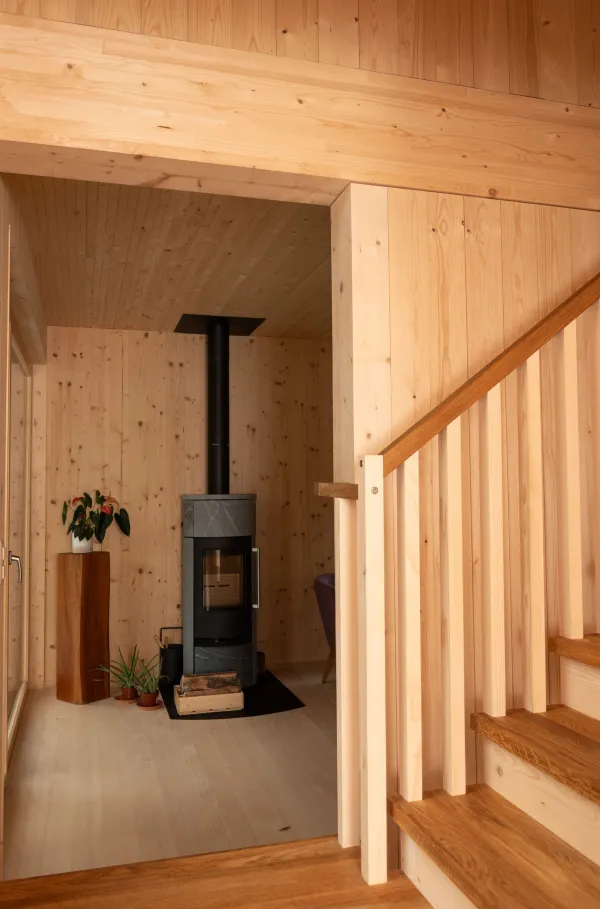 Heller Wohn- und Essbereich in einem Holzhaus mit offener Küche und Kücheninsel. Ein Esstisch mit grünen Stühlen steht vor grossen Fenstern, die auf eine Hanglage blicken. Holzdecken, Holztreppe und warme Beleuchtung prägen den natürlichen Innenraum.