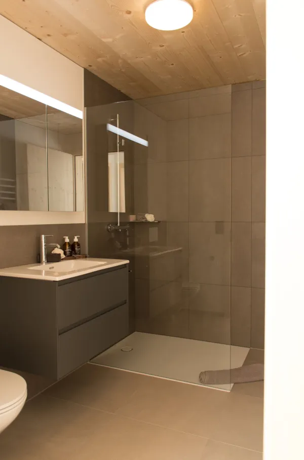 Innenansicht eines modernen Dachgeschosses in Holzbauweise mit sichtbarer Holzdecke und Dachschrägen. Links befindet sich eine dunkle, minimalistische Küche mit Kochinsel, rechts führen grosse Schiebetüren aus Holz und Glas auf einen Balkon. Der helle Holzboden und das einfallende Tageslicht schaffen eine warme, offene Wohnatmosphäre.