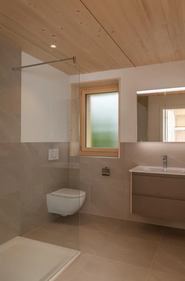 Innenansicht eines hellen Wohnraums in Holzbauweise mit sichtbarer Holzdecke und Holzboden. Grosse, bodentiefe Schiebetüren aus Glas führen auf einen überdachten Balkon mit Holzgeländer. Durch die Fenster fällt viel Tageslicht ein und ermöglicht den Blick nach draussen.