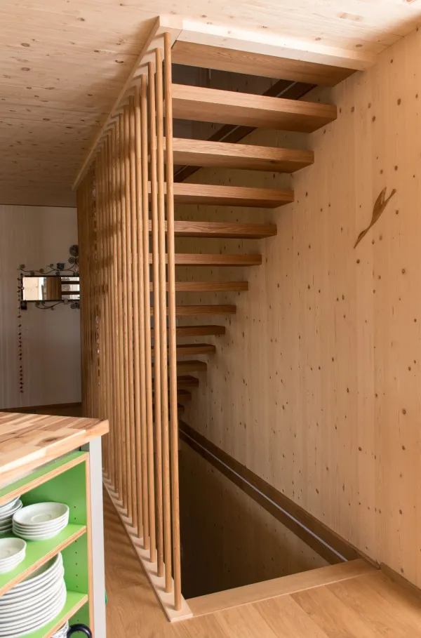 Innenansicht eines offenen Wohn- und Essbereichs in Holzbauweise. Decke und Wände sind mit hellem Holz verkleidet. In der Mitte steht eine Kücheninsel mit offenem Regal und Geschirr. Im Hintergrund befinden sich ein Esstisch mit Stühlen, ein Sitzbereich sowie grosse Fenster, die viel Tageslicht in den wohnlichen Raum lassen.