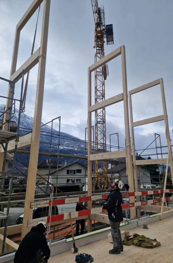 Zimmerleute arbeiten an der Holzrahmenkonstruktion eines mehrgeschossigen Gebäudes auf einer Baustelle. Die tragenden Wände aus Holz werden vor Ort montiert, während ein Baukran über dem Bauplatz steht. Im Hintergrund erstrecken sich Dorfhäuser und eine beeindruckende alpine Berglandschaft