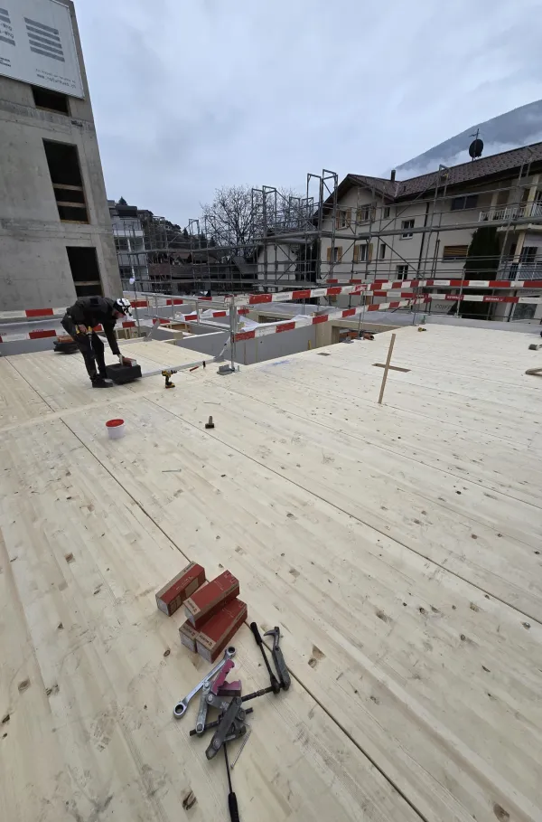 Zimmerleute arbeiten an der Holzrahmenkonstruktion eines mehrgeschossigen Gebäudes auf einer Baustelle. Die tragenden Wände aus Holz werden vor Ort montiert, während ein Baukran über dem Bauplatz steht. Im Hintergrund erstrecken sich Dorfhäuser und eine beeindruckende alpine Berglandschaft