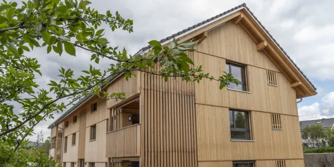 Aussenansicht eines neu errichteten Mehrfamilienhauses in moderner Holzbauweise mit senkrechter Holzfassade und Satteldach. Balkone und Fassadenelemente sind aus Holz gestaltet. Im Vordergrund sind Pflanzen und Baustellenmaterialien zu sehen, im Hintergrund ein Wohnquartier unter bewölktem Himmel.