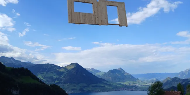 Vorgefertigtes Holzwandelement mit Fenster- und Türöffnung wird von einem Kran über einer alpinen Berglandschaft transportiert. Im Hintergrund sind grüne Berghänge und ein See zu sehen, welche die präzise Montage von Holzbauelementen in anspruchsvollem Gelände veranschaulichen.