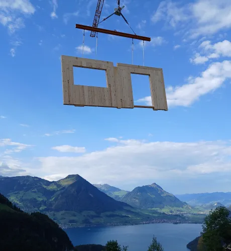 Vorgefertigtes Holzwandelement mit Fenster- und Türöffnung wird von einem Kran über einer alpinen Berglandschaft transportiert. Im Hintergrund sind grüne Berghänge und ein See zu sehen, welche die präzise Montage von Holzbauelementen in anspruchsvollem Gelände veranschaulichen.