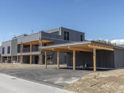 Mehrfamilienhaus Truberholzhaus Recherswil, Schweizer Holz, Truberholzwände sind nicht sichtbar, die Dübel sind nicht sichtbar, Fassade ist vorvergraut, es hat eine Attikawohnung, im Erdgeschoss und Obergeschoss hat es Loggaeinschnitte (Terrasse, Balkon), moderner Neubau, auf dem Dach ist ein Sonnensegel, Flachdach mit Solarpanel, es hat kein Keller dafür ein zusätzliches Gebäude mit Autounterstand und Lagerraum, Umschwung mit Rasen 