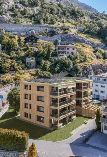 Visualisierung eines modernen Mehrfamilienhauses in Holzbauweise mit grossen Balkonen und begrüntem Dach mit Solaranlage. Das Gebäude fügt sich in eine alpine Dorflandschaft mit traditionellen Häusern, Strassen und einem bewaldeten Berghang im Hintergrund ein