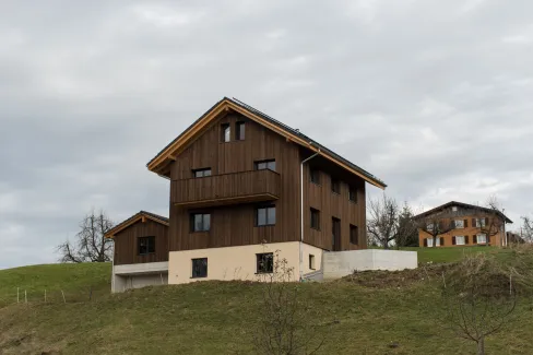 Neubau eines modernen Holzhauses am Hang mit dunkler Holzfassade, Balkon und grosser Fensterfront. Das Gebäude steht in ländlicher Umgebung mit Wiese und weiteren Häusern im Hintergrund unter bewölktem Himmel.