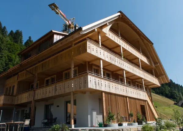 Mehrstöckiges Holzhaus im traditionellen Schweizer Stil mit kunstvoll verzierten Holzbalkonen und grosszügigem Dachüberstand. Die Fassade besteht aus hellem, massivem Holz, umgeben von einer grünen Hanglandschaft bei klarem Himmel.