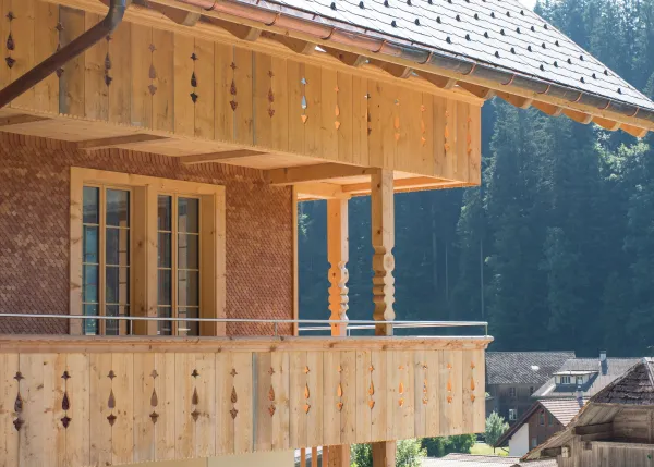 Mehrstöckiges Holzhaus im traditionellen Schweizer Stil mit kunstvoll verzierten Holzbalkonen und grosszügigem Dachüberstand. Die Fassade besteht aus hellem, massivem Holz, umgeben von einer grünen Hanglandschaft bei klarem Himmel.
