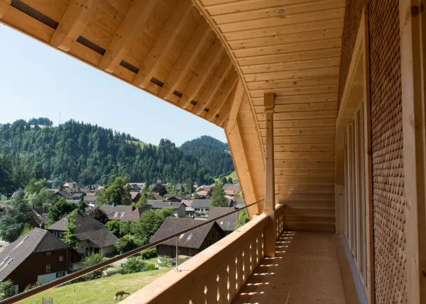 Mehrstöckiges Holzhaus im traditionellen Schweizer Stil mit kunstvoll verzierten Holzbalkonen und grosszügigem Dachüberstand. Die Fassade besteht aus hellem, massivem Holz, umgeben von einer grünen Hanglandschaft bei klarem Himmel.