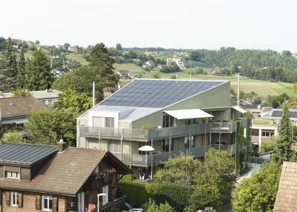 Aussenansicht eines modernen, mehrstöckigen Wohngebäudes in Holzbauweise mit hellgrüner Fassade. Das Haus verfügt über grosse Balkone mit Metallgeländern und Kletterpflanzen, die vertikal an den Fassadenelementen emporwachsen. Vor dem Gebäude liegt ein gepflegter Garten mit jungen Bäumen, Sträuchern und geschwungenen Wegen. Die Architektur wirkt offen und einladend, mit vielen Fenstern und einer klar strukturierten Formensprache. Im Vordergrund sind ein Kiesweg und ein Zaun sichtbar, während der Himmel leic