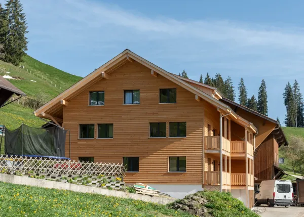 Aussenansicht eines neu errichteten Wohnhauses in Holzbauweise mit Satteldach und Balkonen, eingebettet in eine hügelige, ländliche Landschaft. Das Gebäude steht neben bestehenden landwirtschaftlichen Bauten. Im Vordergrund eine Wiese, im Hintergrund ein begrünter Hang mit Bäumen unter blauem Himmel.