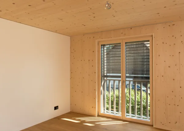 Innenansicht eines modernen Dachgeschosses in Holzbauweise mit sichtbarer Holzdecke und Dachschrägen. Links befindet sich eine dunkle, minimalistische Küche mit Kochinsel, rechts führen grosse Schiebetüren aus Holz und Glas auf einen Balkon. Der helle Holzboden und das einfallende Tageslicht schaffen eine warme, offene Wohnatmosphäre.