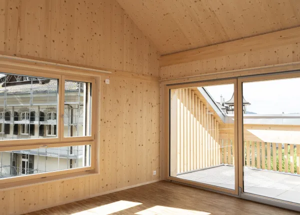 Innenansicht eines hellen Wohnraums in Holzbauweise mit sichtbarer Holzdecke und Holzboden. Grosse, bodentiefe Schiebetüren aus Glas führen auf einen überdachten Balkon mit Holzgeländer. Durch die Fenster fällt viel Tageslicht ein und ermöglicht den Blick nach draussen.