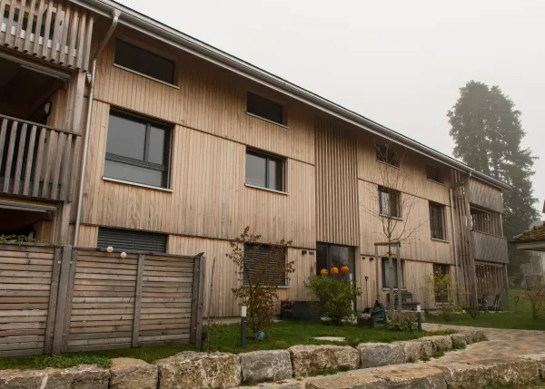 Aussenansicht eines neu errichteten Mehrfamilienhauses in moderner Holzbauweise mit senkrechter Holzfassade und Satteldach. Balkone und Fassadenelemente sind aus Holz gestaltet. Im Vordergrund sind Pflanzen und Baustellenmaterialien zu sehen, im Hintergrund ein Wohnquartier unter bewölktem Himmel.