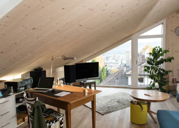 Innenansicht eines offenen Wohn- und Essbereichs in Holzbauweise. Decke und Wände sind mit hellem Holz verkleidet. In der Mitte steht eine Kücheninsel mit offenem Regal und Geschirr. Im Hintergrund befinden sich ein Esstisch mit Stühlen, ein Sitzbereich sowie grosse Fenster, die viel Tageslicht in den wohnlichen Raum lassen.